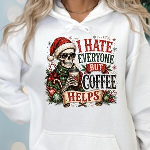 May include: A white hooded sweatshirt featuring a graphic of a skeleton wearing a Santa hat, sunglasses, and a plaid scarf while holding a coffee cup. The text reads I HATE EVERYONE BUT COFFEE HELPS, surrounded by festive holiday decorations.