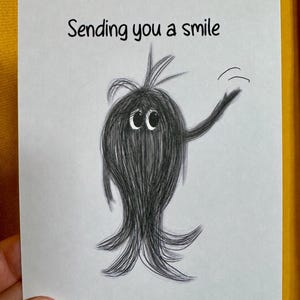 May include: A white greeting card featuring a black pencil-style sketch of a fuzzy, round creature with large eyes and a tuft of hair on top. The creature is waving one arm, and the text "Sending you a smile" is printed in black above it.