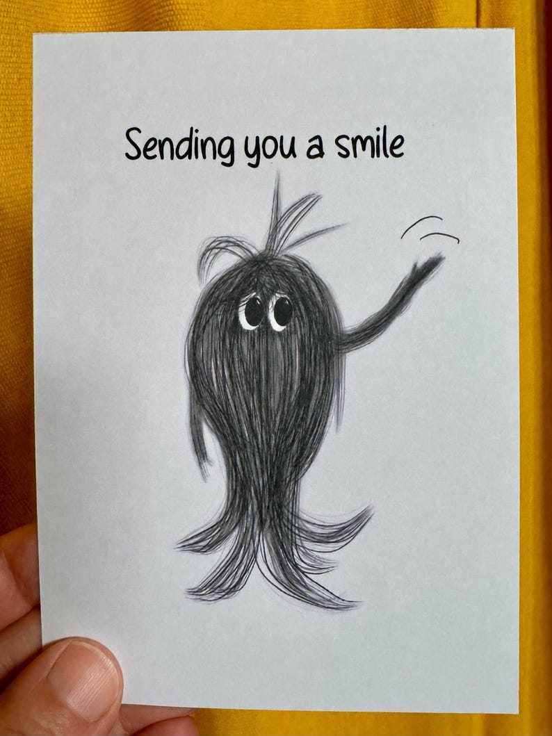 May include: A white greeting card featuring a black pencil-style sketch of a fuzzy, round creature with large eyes and a tuft of hair on top. The creature is waving one arm, and the text "Sending you a smile" is printed in black above it.