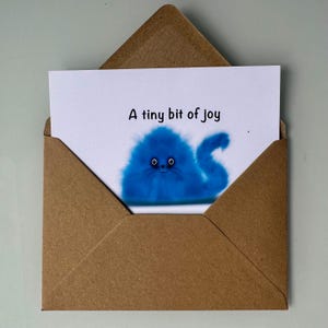 Op de afbeelding: Een wenskaart met de tekst "A tiny bit of joy" in een bruine envelop. De kaart toont een pluizig blauw wezen met een staart. De envelop is open en toont de kaart.
