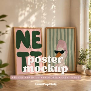 Könnte beinhalten: Zwei gerahmte Poster in Holzrahmen. Das linke Poster zeigt den großen dunkelgrünen Schriftzug NETT HIER auf hellrosa Hintergrund. Das rechte Poster zeigt eine Illustration einer Ente mit rosa Handtuchwickel und schwarzer Sonnenbrille.