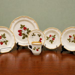 Puede incluir: Un juego de vajilla de cerámica con base color crema y borde amarillo festoneado. Cada pieza está decorada con una ilustración botánica de fresas rojas, hojas verdes y flores blancas. El juego incluye platos, un platillo y una taza.