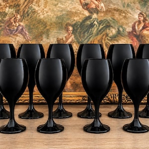 May include: A set of nine solid black glass wine glasses with rounded bowls and slender stems. Each glass features a smooth, opaque finish and a wide circular base, arranged in a grid formation on a light wood surface for a uniform appearance.