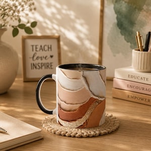 Könnte beinhalten: Eine Keramik-Kaffeetasse mit schwarzem Henkel und Rand, mit einem geschichteten abstrakten Design in Beige-, Braun- und Weißtönen. Die Tasse steht auf einem beigefarbenen Untersetzer. Ein Schild liest "Teach love Inspire".