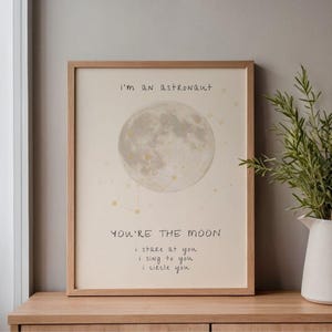 May include: A framed print with a light beige background features a full moon illustration and constellations. The text reads, "I'm an astronaut" and "You're the moon, I stare at you, I sing to you, I circle you." The frame is made of light brown wood.