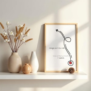 May include: A framed print with the text "change your zip code turns out that you're still an asshole." The print is in a light wood frame. There are vases and decorative flowers on a white shelf.