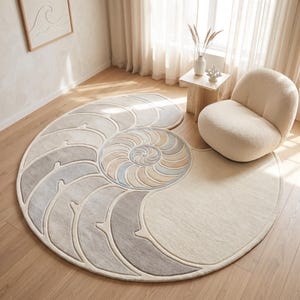 Op de afbeelding: Een groot, rond vloerkleed met een gestileerd nautilusschelpontwerp in tinten crème, beige, lichtblauw en grijs. Het kleed heeft een getufte textuur met uitgesneden lijnen die de spiraalvormige segmenten van het schelppatroon op het oppervlak definiëren.