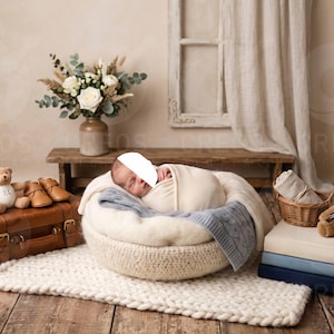 May include: A newborn infant wrapped in a cream-colored swaddle rests in a round, cream-colored knit basket. The scene includes a brown suitcase, two teddy bears, a bouquet of white flowers, a stack of three books, and a small woven basket.