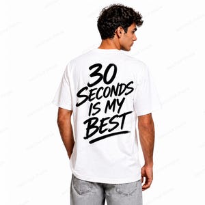 Può includere: T-shirt bianco con la scritta "30 SECONDS IS MY BEST" in nero, in grassetto, con un carattere in stile manoscritto. La maglietta è un classico girocollo. La persona indossa jeans grigi.