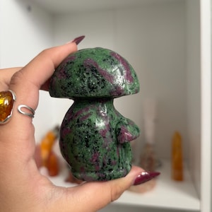 May include: A carved mushroom figurine made of polished zoisite with ruby inclusions. The stone features a mottled green base colour with dark black speckles and vibrant magenta-red patches distributed across the cap and the rounded stem.