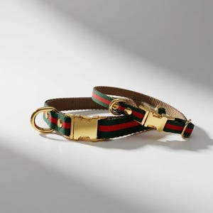 May include: Two pet collars featuring a green and red striped webbing strap with gold-toned metal side-release buckles and D-rings. The collars are constructed with a durable fabric material and include adjustable metal sliders for a secure fit.