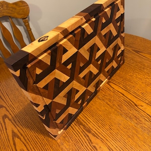 May include: A handcrafted wooden cutting board with a geometric pattern. The board features a mosaic design of various wood tones, including light, medium, and dark browns. The edges are a darker brown. A small logo is visible on the side.
