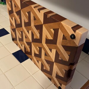May include: A rectangular wooden cutting board with a geometric pattern. The board is made of various wood tones, including light, medium, and dark browns. It has a handle on one side and measures approximately 18 inches long and 12 inches wide.