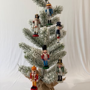Puede incluir: Un pequeño árbol de Navidad artificial flocado con una base envuelta en arpillera, decorado con seis adornos colgantes de cascanueces de madera. Los cascanueces lucen uniformes variados en rojo, azul, blanco, verde y dorado con sombreros.