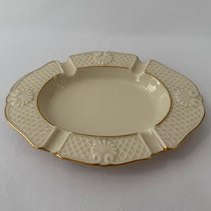 May include: An oval cream-coloured ceramic ashtray featuring a textured lattice pattern on the rim, accented with four embossed shell motifs and a thin gold-painted trim along the edges and the central basin of the dish.