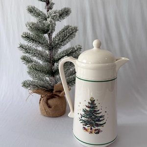 May include: A cream-colored ceramic coffee pot with a lid and handle, featuring a green band around the top and bottom and a central illustration of a decorated Christmas tree surrounded by colorful gift boxes and small gold stars.