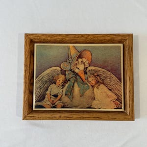 May include: A rectangular wooden frame containing a print of a large white goose wearing a blue bonnet and a polka-dot bow, flanked by two children with white wings. The illustration features soft pastel colors and a muted blue background.