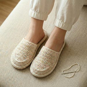 Puede incluir: Un par de zapatillas de crochet beige hechas a mano. Las zapatillas tienen un diseño de punta redondeada y un patrón de puntada texturizada. La imagen muestra las zapatillas sobre una superficie de color neutro, sugiriendo comodidad y uso doméstico.