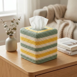 Può includere: Copertura per scatola di fazzoletti quadrata in filato all'uncinetto. La copertura presenta strisce orizzontali verdi, gialle e bianco sporco. Fazzoletti bianchi visibili in cima. Posizionata su un tavolo di legno.