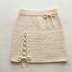 May include: Cream-colored crochet skirt with a lace-up detail on the side and a bow at the waist. The skirt has a ribbed waistband and a straight silhouette. The crochet work is tight and even, creating a textured look.