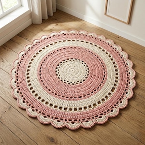 May include: A round, handmade crochet rug in a spiral design. The rug features alternating bands of light pink and cream colors, with a scalloped edge. The rug is placed on a hardwood floor, near a window.