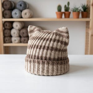 Puede incluir: Un gorro hecho a mano con orejas de gato en tonos marrones y beige. El gorro de punto acanalado presenta un patrón de rayas y orejas puntiagudas en la parte superior. El gorro se muestra sobre una superficie blanca.