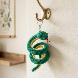 May include: A crocheted snake keychain featuring a coiled body with a vibrant green top and a pale yellow underside. The snake has small black bead eyes and a red yarn tongue, attached to a silver-toned metal split ring for hanging purposes.