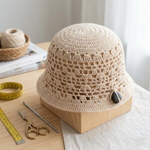 Peut inclure: Un chapeau seau en crochet beige clair avec un motif ajouré. Le chapeau est présenté sur une forme en bois, avec un accent en pierre sombre. Un mètre ruban, des ciseaux et un crochet sont visibles.