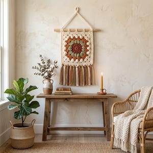 May include: A cream-colored, square crochet wall hanging with a geometric design in shades of orange, green, and brown. The wall hanging is suspended from a wooden dowel and features long, colorful tassels. The scene includes a wooden console table, a potted plant, and a wicker chair.