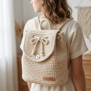 May include: Cream-colored crochet backpack with a flap closure and drawstring. The bag features adjustable straps and a small handle. A leather tag on the front reads "HANDCRAFTED EVERYDAY BACKPACK".