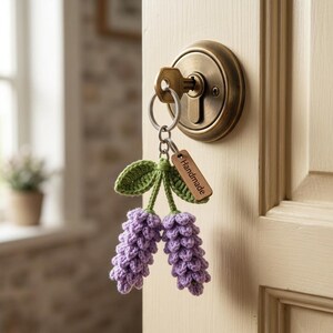 Puede incluir: Un llavero de ganchillo hecho a mano con dos racimos de flores moradas con hojas verdes. El llavero está sujeto a una llave en una cerradura de latón. También hay una pequeña etiqueta de madera con la palabra "Handmade".