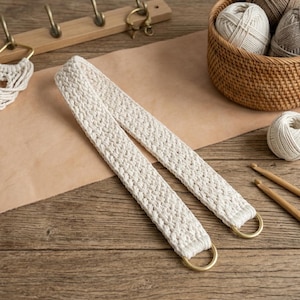 Puede incluir: Correa de macramé color crema con anillos de latón, sobre una superficie de madera. La correa está hecha de algodón tejido y forma parte de un proyecto de artesanía. Otros suministros de artesanía son visibles en el fondo, incluyendo hilo y ganchos.