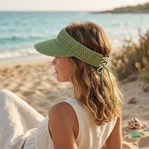 Op de afbeelding: Een groene zonnevizier met een geweven rand en een gehaakte band. De zonnevizier heeft een striksluiting aan de achterkant en wordt door een persoon op het strand gedragen. De zonnevizier is lichtgroen van kleur.