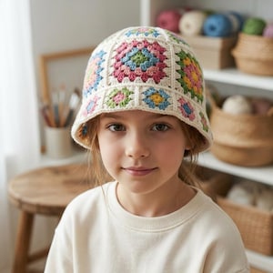 May include: A cream-coloured crochet bucket hat with colourful floral squares. The hat has a brim and is worn by a person. The squares are in shades of pink, blue, green, and yellow. The background includes shelves with yarn and baskets.