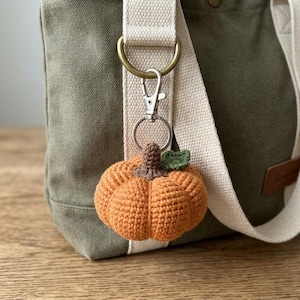 May include: A crocheted pumpkin keychain in shades of orange, brown, and green, attached to a silver-colored metal clasp and ring. The pumpkin is hanging from a canvas bag with a cream-colored strap. The background is a wooden surface.