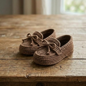 May include: A pair of brown crochet baby shoes with a loafer design. Each shoe features a decorative bow on the top. The shoes are made of a textured yarn and sit on a wooden surface.