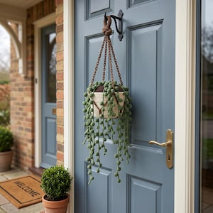 Peut inclure: Une plante suspendue dans un pot en crochet crème avec un bord et une chaîne marron. La plante a de longues tiges vertes pendantes avec de petites feuilles rondes. Le pot est suspendu à un crochet en métal noir sur une porte bleue.