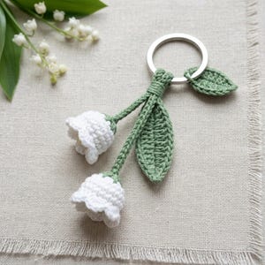 May include: A handmade crochet keychain featuring two white lily of the valley flowers with green stems and leaves. The flowers are attached to a silver-colored key ring. The keychain is set against a neutral linen background.