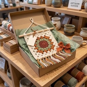 May include: A cream-colored, square crochet wall hanging with a floral design in shades of orange, green, and brown, displayed in a cardboard box. The hanging has a wooden dowel and a tassel fringe. A blank tag hangs from the dowel. A rust-colored bow is attached.