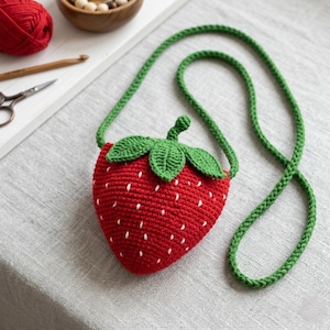 Può includere: Una borsetta a forma di fragola fatta a mano, all'uncinetto in rosso, con gambo e foglie verdi. La borsa ha un cinturino verde e dettagli di semi bianchi. Un accessorio stravagante.