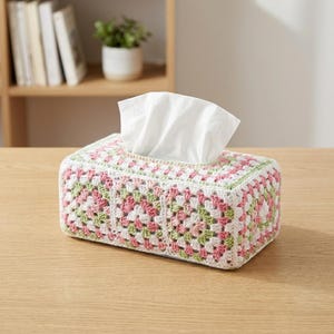 Puede incluir: Funda rectangular para caja de pañuelos de ganchillo. El diseño presenta cuadrados de la abuela en blanco, rosa y verde. Los pañuelos blancos sobresalen de la abertura superior. Colocada sobre una superficie de madera.