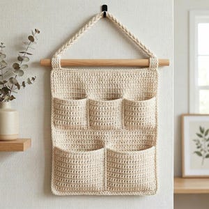 May include: A cream-colored, crocheted wall organizer with six pockets. It hangs from a wooden dowel and a braided loop. The neutral tones and handmade texture create a cozy, rustic aesthetic, suitable for home organization.