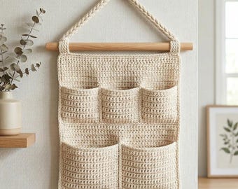 May include: A cream-colored, crocheted wall organizer with six pockets. It hangs from a wooden dowel and a braided loop. The neutral tones and handmade texture create a cozy, rustic aesthetic, suitable for home organization.