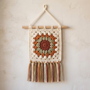 May include: A cream-colored, square crochet wall hanging with a circular, multi-colored design in the center. The design features shades of brown, orange, and green. The hanging is suspended from a wooden dowel with a cream-colored rope and has a fringe of tassels.
