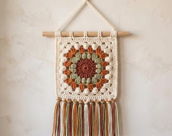 May include: A cream-colored, square crochet wall hanging with a circular, multi-colored design in the center. The design features shades of brown, orange, and green. The hanging is suspended from a wooden dowel with a cream-colored rope and has a fringe of tassels.