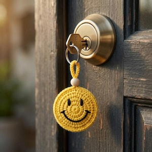 Op de afbeelding: Een gele gehaakte smiley-sleutelhanger met een zwart gestikte mond en ogen, hangend aan een sleutel in een messing deurslot. De sleutelhanger heeft een houten kraal en een lus om op te hangen.