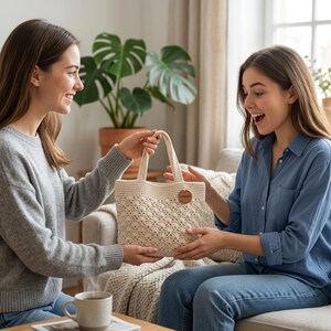 May include: A cream-colored, crocheted tote bag with a diamond pattern is being presented. The bag has two handles and a small, round tag with text. The bag is held by two people. A cup of coffee sits on a table in the foreground.