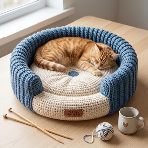 Può includere: Un letto per animali domestici all'uncinetto con un gatto tabby arancione che dorme. Il letto è rotondo, con un centro color crema e un bordo blu a coste. Una piccola etichetta recita "PET BED PATTERN". Ferri da maglia, filo e una tazza sono nelle vicinanze.