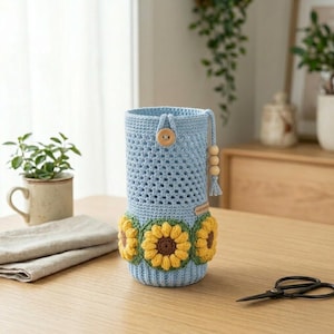 May include: A light blue crochet water bottle holder with a sunflower design. The holder has a wooden button closure and a drawstring with wooden beads. The bottom of the holder is decorated with yellow and brown sunflowers.