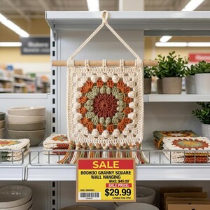 May include: A square, cream-colored, crocheted wall hanging with a colorful circular design in shades of orange, green, and brown. The wall hanging is suspended from a wooden dowel and has a fringe at the bottom. A sale sign is visible.